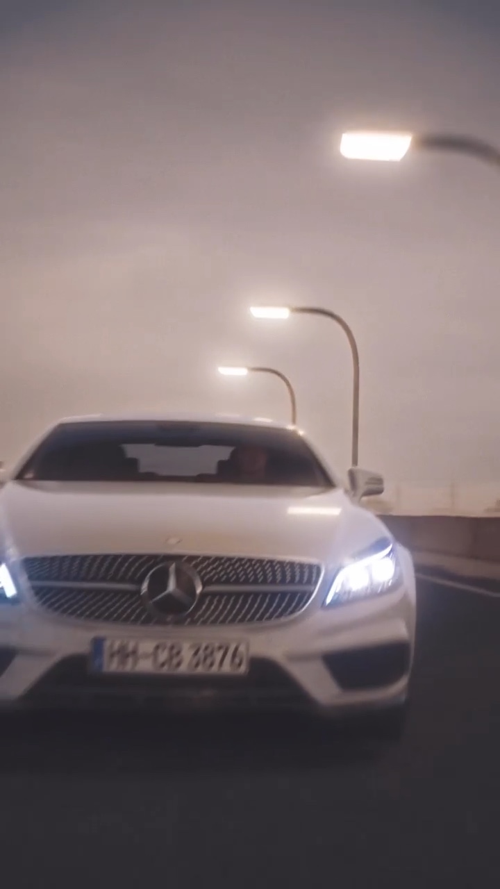 Front view of a silver Mercedes CLS with headlights on, on a road at dusk, street lights in the background